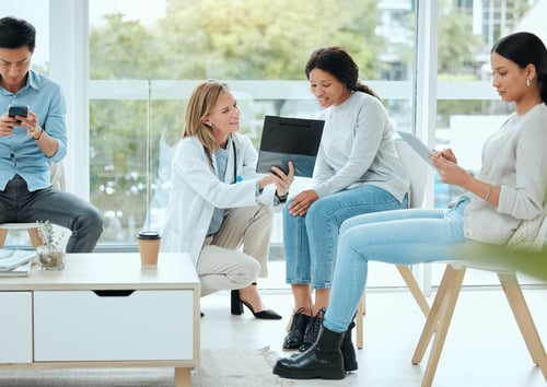 A doctor in a white coat shows a tablet to a smiling woman in a modern room with large windows.