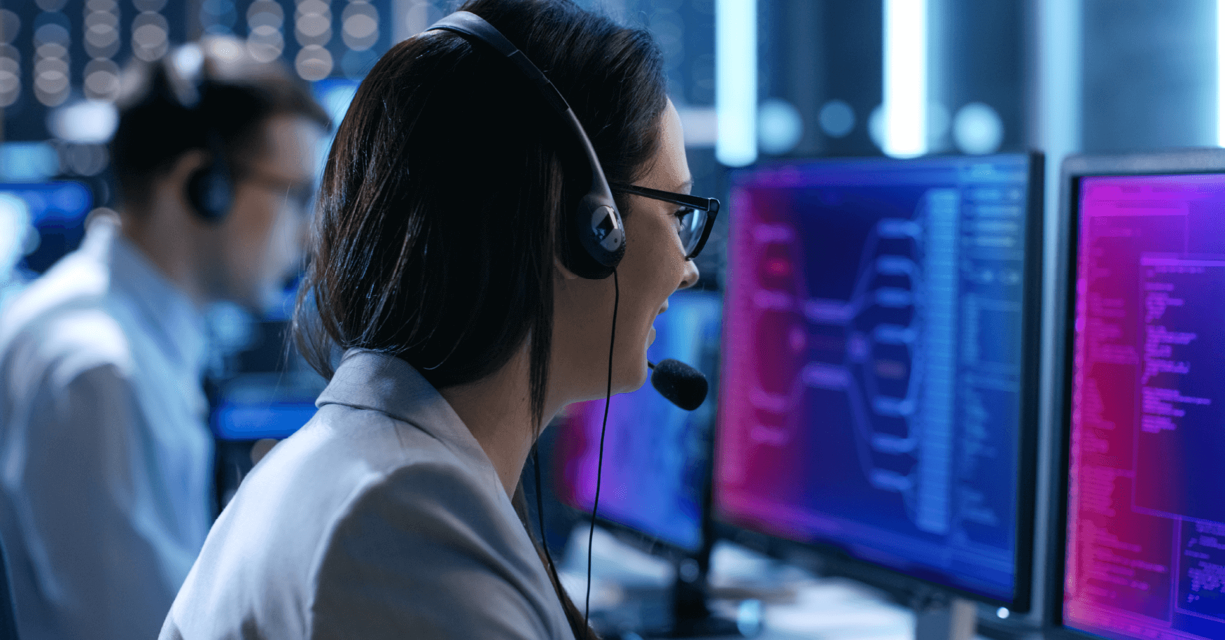 Professional woman wearing headset working at computer workstation with multiple monitors displaying data in modern office environment